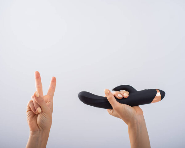 Faceless woman holding black dildo and showing peace sign on white background.