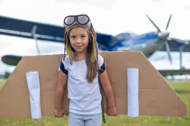 Karton kanatlı pilot kostümlü küçük bir kız uçağın arka planına karşı çimlerde koşuyor. Şapkalı ve gözlüklü bir çocuk uçmayı hayal eder..