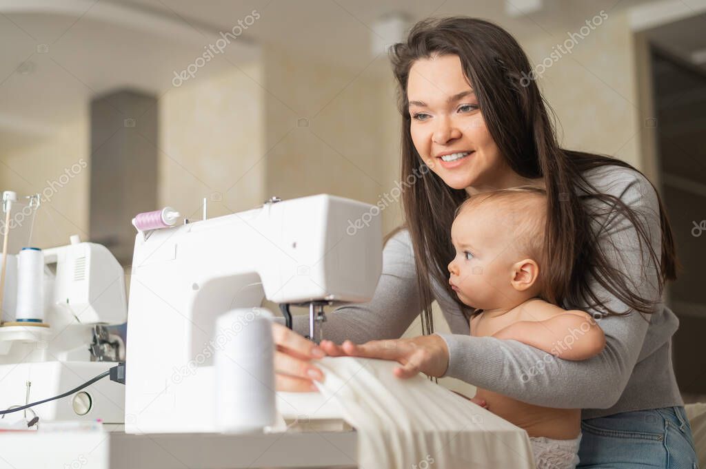 Young woman sews at home and holds a small child. Mom teaches her little son to sew on a sewing machine