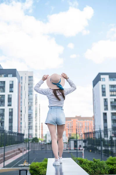 Feliz mujer joven caucásica en un sombrero y pantalones cortos camina en una calle de la ciudad en un día caluroso de verano —  Fotos de Stock