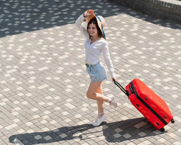 Glückliche kaukasische junge Frau mit Hut und kurzen Hosen, die in offenem Alter einen großen roten Koffer hält — Stockfoto