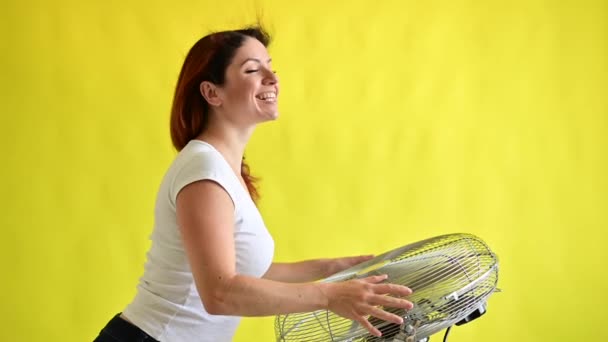 Une belle femme aux cheveux roux est refroidie debout sur un grand ventilateur électrique sur un fond jaune. Fille avec les cheveux se développant dans le vent. Dispositif de refroidissement de l'air. — Video