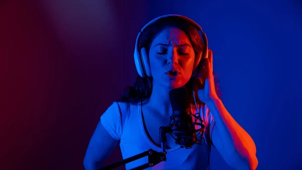 Mujer caucásica en auriculares cantando en un micrófono con luz de neón sobre un fondo negro. Una chica emocional está grabando una canción en un estudio de grabación —  Fotos de Stock