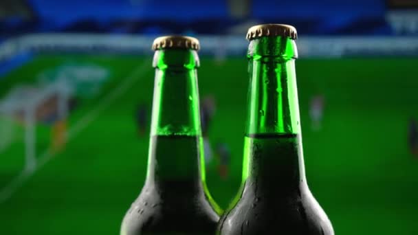 サッカー選手権放送を背景に、 3本のグリーングラスビールボトルが回転する — ストック動画
