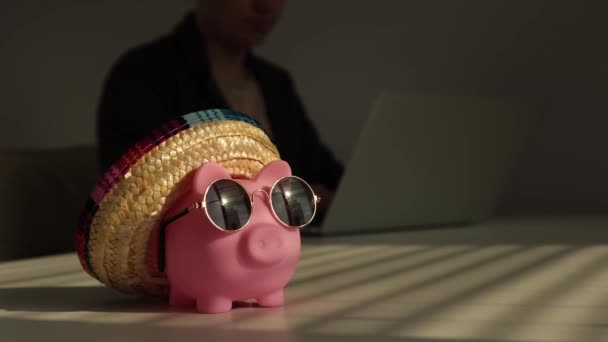 Piggy bank w okularach przeciwsłonecznych i sombrero w tle kobieta pracuje dla laptopa — Wideo stockowe