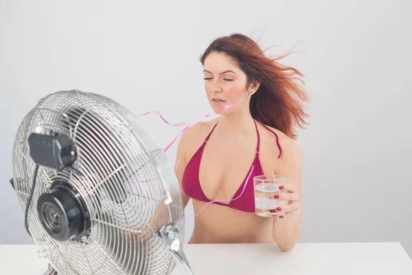 Mujer sonriente pelirroja en bikini bebe una bebida fría y disfruta del viento que sopla de un ventilador eléctrico sobre un fondo blanco. Control climático en un caluroso día de verano — Foto de Stock