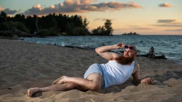 Divertente uomo calvo con la barba rossa vestita in biancheria intima in posa sulla spiaggia. Una parodia maschile divertente di una ragazza glamour. — Foto Stock