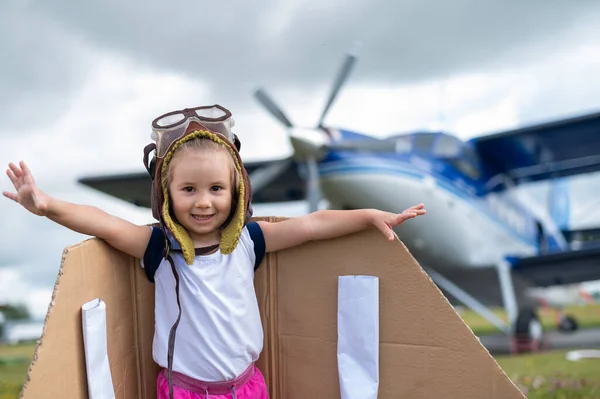 Ein nettes kleines Mädchen mit Mütze und Pilotenbrille auf dem Hintergrund eines Flugzeugs. Das Kind träumt davon, Pilot zu werden. — Stockfoto