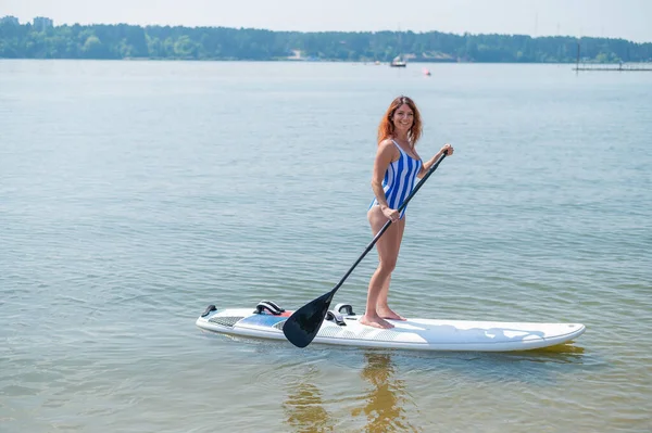 Kaukasische Frau im gestreiften Badeanzug fährt auf einem SUP-Board. Das Mädchen bevorzugt aktive Erholung. — Stockfoto