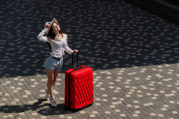 Glückliche kaukasische junge Frau in Hut und Shorts mit einem großen roten Koffer im Freien. — Stockfoto