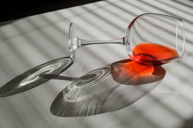 Bir kadeh kırmızı şarap yan yatar beyaz bir masada, panjurların gölgesinde.