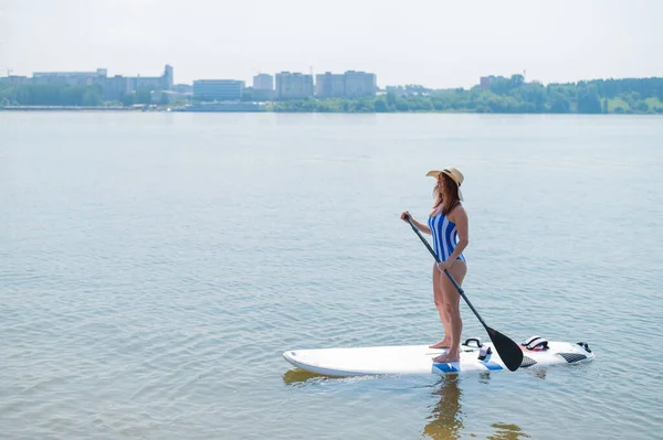 Eine rothaarige Frau in gestreiftem Badeanzug und Strohhut schwimmt auf einem Paddelbrett auf dem See — Stockfoto