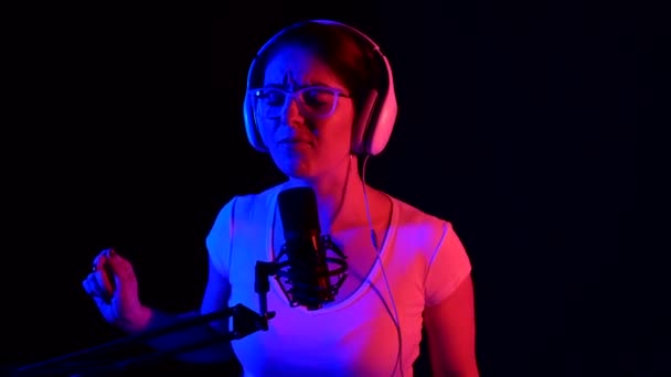 Blanke vrouw met bril en koptelefoon zingt in een microfoon in neon licht op een zwarte achtergrond. Een emotioneel meisje neemt een lied op in een opnamestudio. — Stockvideo