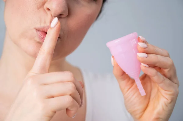 Retrato de cerca de una mujer sosteniendo una copa menstrual rosa sobre un fondo blanco. Chica sosteniendo el dedo índice en su boca haciendo una señal de silencio. Tímido sobre la menstruación — Foto de Stock