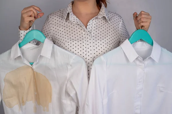 A woman compares two white shirts before and after washing. The girl is holding one blouse, clean and ironed, and the other, dirty with coffee stains