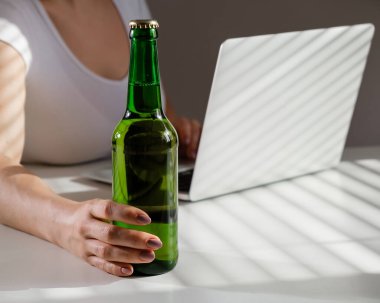 Beyaz kadın dizüstü bilgisayarda çalışıyor ve cam şişeden bira içiyor.