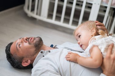 Kafkasyalı adam sırt üstü uzanıyor ve kızını çocuk odasında onun üstünde tutuyor.