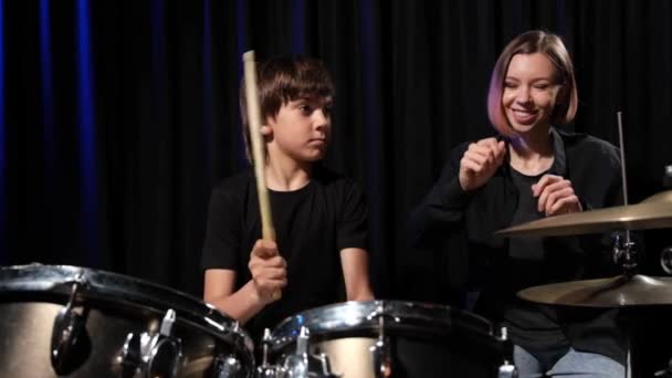 Joven mujer caucásica enseña a un niño a tocar la batería en el estudio sobre un fondo negro. Estudiante de música — Vídeos de Stock