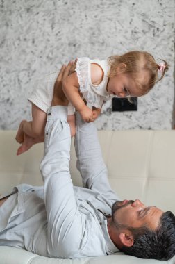 Beyaz bir adam kanepede uzanıyor ve küçük bir kızı üzerinde tutuyor. Şefkatli bir baba çocuğu büyüttü. Mutlu babalar.