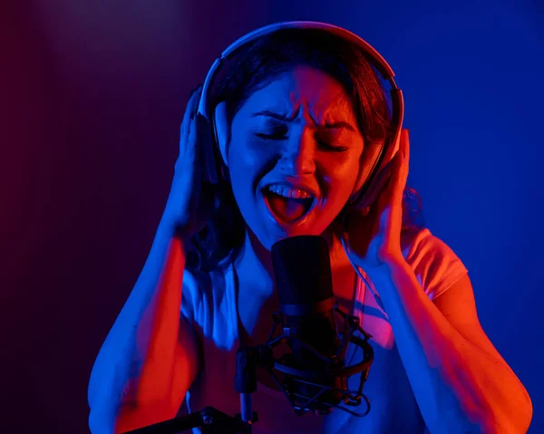 Fehér nő fülhallgatóban, aki neon fényben énekel egy mikrofonba, fekete háttérrel. Egy érzelmes lány felvesz egy dalt egy stúdióban. — Stock Fotó