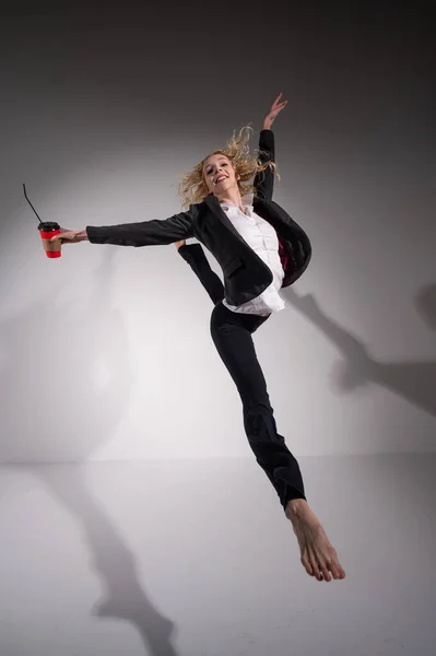 Χαριτωμένη ξυπόλητη μπαλαρίνα σε ένα επαγγελματικό κοστούμι πηδώντας με ένα ποτήρι καφέ στα χέρια της σε λευκό φόντο. — Φωτογραφία Αρχείου