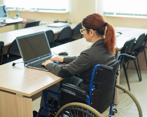 Młoda kobieta pracuje na laptopie siedząc na wózku inwalidzkim w sali wykładowej. Warunki nauczania osoby niepełnosprawnej — Zdjęcie stockowe