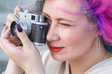 Beyaz saçlı, kıvırcık saçlı bir kadın eski bir retro kamerayla dışarıda fotoğraf çekiyor.