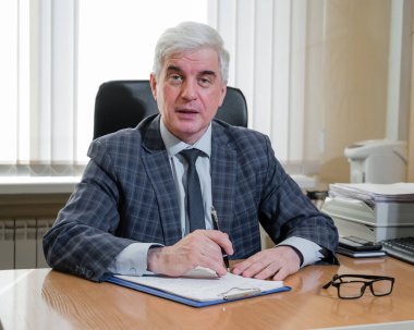 Kendinden emin, gri saçlı, ekose takım elbiseli adam ofisinde masasında oturuyor.