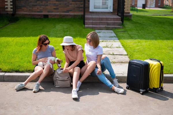 Drei kaukasische Frauen und ein Hund gehen auf Reisen. Die Mädchen sitzen mit Koffern auf dem Bordstein und warten auf ein Taxi. Sommerferienkonzept mit Freundinnen — Stockfoto