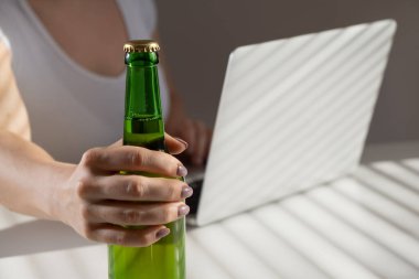 Beyaz kadın dizüstü bilgisayarda çalışıyor ve cam şişeden bira içiyor.