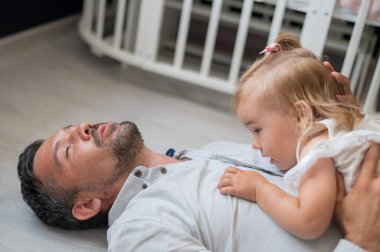 Kafkasyalı adam sırt üstü uzanıyor ve kızını çocuk odasında onun üstünde tutuyor.