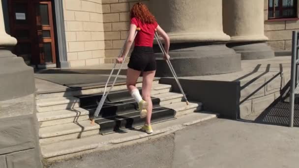 Junge rothaarige Frau mit Knöchelverletzung steigt auf Krücken Treppe hinauf. — Stockvideo