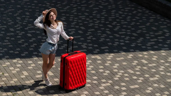 Feliz caucasiano jovem mulher em chapéu e shorts segurando uma grande mala vermelha ao ar livre. — Fotografia de Stock