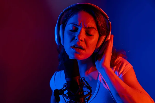 Mulher branca em fones de ouvido cantando em um microfone em luz de néon em um fundo preto. Uma garota emocional está gravando uma música em um estúdio de gravação — Fotografia de Stock