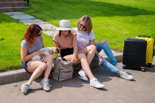 Tres mujeres caucásicas y un perro se van de viaje. Las chicas están sentadas en la acera con maletas y esperando un taxi. Concepto de vacaciones de verano junto con novias —  Fotos de Stock