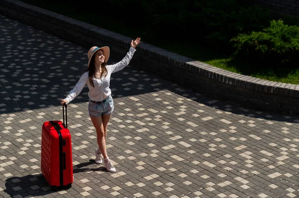 Glückliche kaukasische junge Frau in Hut und Shorts mit einem großen roten Koffer im Freien. — Stockfoto