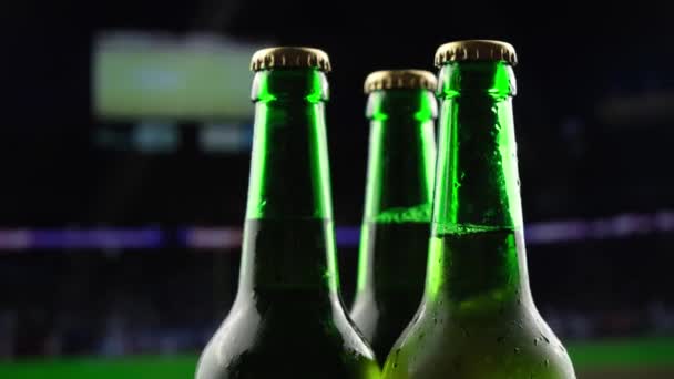 Tres botellas de vidrio verde de cerveza giran en la oscuridad contra el telón de fondo de una emisión de un partido de fútbol americano — Vídeo de stock