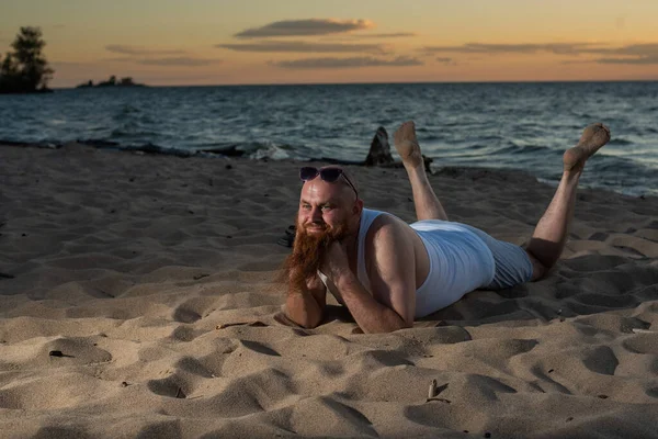 Divertente uomo calvo con la barba rossa in posa sulla spiaggia al tramonto. Una parodia maschile divertente di una ragazza glamour. — Foto Stock