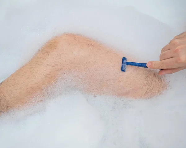 한 남자가 편안 한 목욕을 하고 다리를 면도 하고 있는 이상 한 그림입니다. 거품 목욕을 하고 있는 남성 발의 근접 사진. 위에서 본 풍경. — 스톡 사진