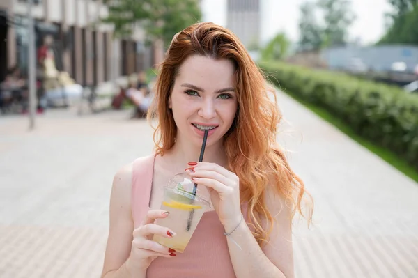 夏天，年轻美丽的红头发女人带着牙套，在户外喝着凉柠檬水。一个带雀斑的微笑女孩的画像. — 图库照片