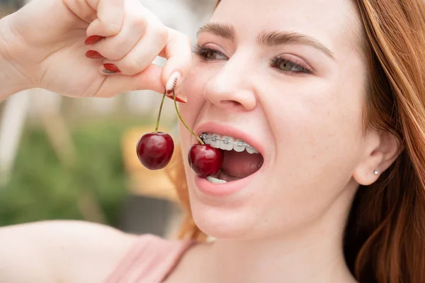 Belle jeune femme aux cheveux roux avec des bretelles sur les dents mange des cerises douces en été à l'extérieur — Photo