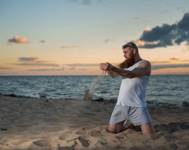 Gün batımında sahilde poz veren kızıl sakallı komik kel adam. Çekici bir kızın esprili erkek parodisi..