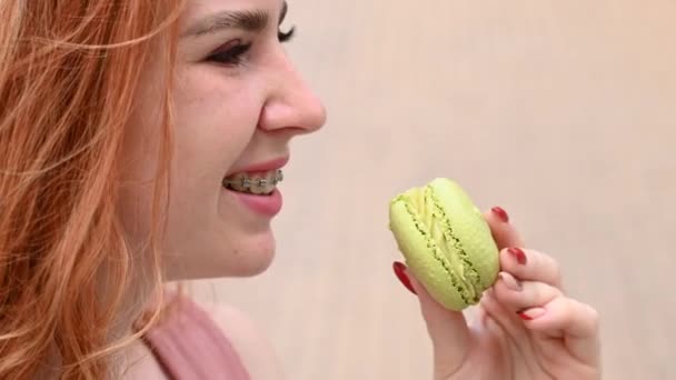 Gyönyörű fiatal nő fogszabályzóval eszik macaroon torta a szabadban — Stock videók