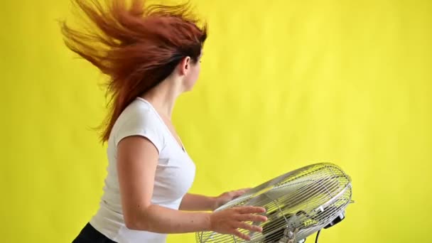 Güzel kızıl saçlı bir kadın, sarı arka planda büyük bir elektrikli vantilatörün başında serinliyor. Saçları rüzgarda savrulan bir kız. Havayı soğutmak için aygıt. — Stok video