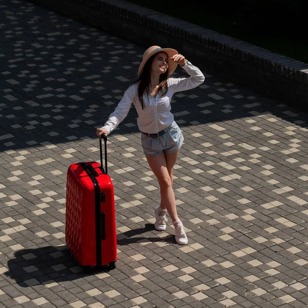 Glückliche kaukasische junge Frau in Hut und Shorts mit einem großen roten Koffer im Freien. — Stockfoto