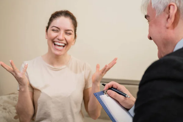 Psicoterapeuta masculino maduro de pelo gris con portapapeles en sesión con paciente femenino. Sonriente mujer caucásica en una cita con un psicólogo. —  Fotos de Stock