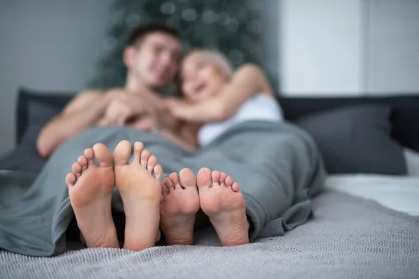 Közelkép a férfi és női lábakról. A szerető pár hazudik és ölelkezik az ágyon egy szürke takaró alatt. Világítószoba. — Stock Fotó