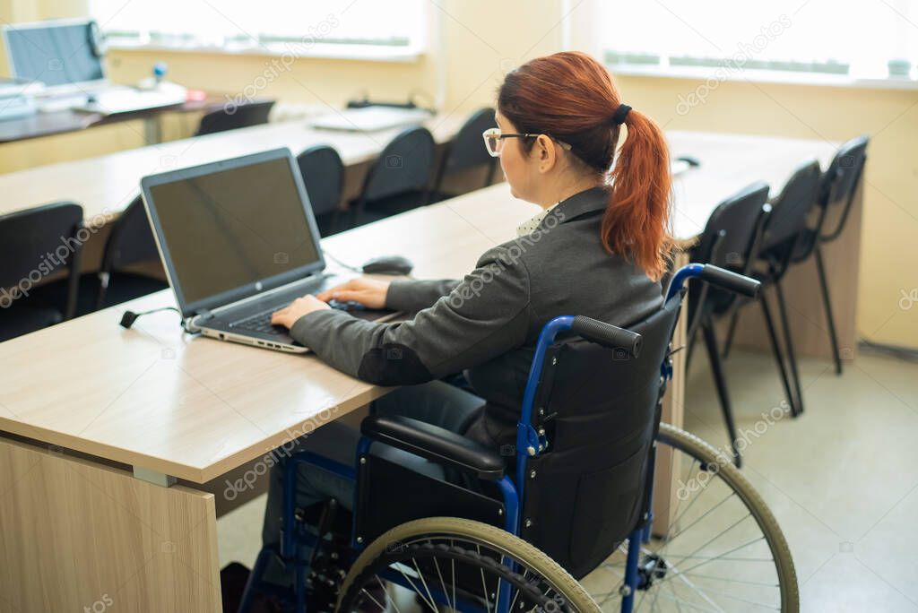 Young woman works at a laptop while sitting in a wheelchair in a university lecture hall. Conditions for teaching a disabled person