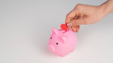 Yüzsüz beyaz kadın küçük bir kalbi beyaz arka planda pembe bir domuz kumbarasına çeviriyor. Aşk tasarruf kavramı
