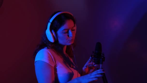 Mujer caucásica en auriculares canta en un micrófono con luz de neón. Una chica emocional está grabando una canción en un estudio de grabación — Vídeos de Stock